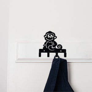 Puede incluir: Perchero de pared de metal negro con un diseño de mono sonriente. El gancho tiene tres ganchos inferiores y una silueta decorativa de mono en la parte superior. Una prenda azul oscuro cuelga de uno de los ganchos. El gancho está montado en una pared blanca.