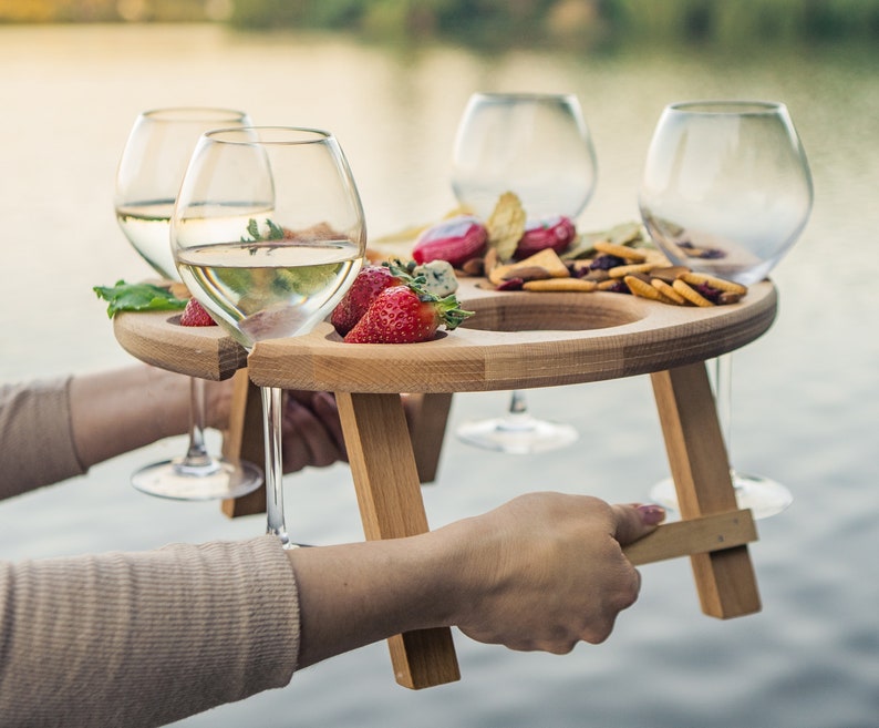 K&ouml;nnte beinhalten: Ein h&ouml;lzerner Picknicktisch mit vier Weinglashaltern und einem Serviertablett. Der Tisch ist aus hellbraunem Holz und hat eine nat&uuml;rliche Oberfl&auml;che. Das Tablett ist gef&uuml;llt mit Snacks, darunter K&auml;se, Cracker und Obst. Der Tisch ist perfekt, um ein Picknick im Freien zu genie&szlig;en.