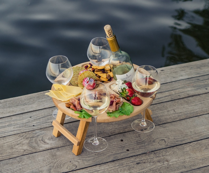 K&ouml;nnte beinhalten: Ein Holz-Picknicktisch mit einem eingebauten Serviertablett. Das Tablett ist gef&uuml;llt mit K&auml;se, Crackern, Obst und anderen Snacks. Auf dem Tisch stehen vier Weingl&auml;ser und eine Flasche Wein.