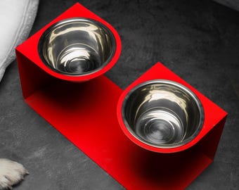May include: A bright red elevated pet feeder stand featuring two circular cutouts that hold two removable stainless steel bowls. The stand has a sleek, modern design with a continuous metal frame that supports both bowls at an elevated height.