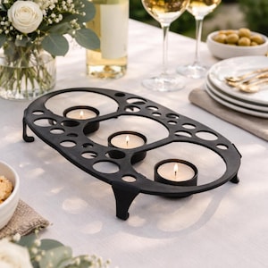 May include: An oval-shaped black metal candle holder featuring a perforated design with various circular cutouts. The frame is supported by four small feet and holds three lit tea light candles in individual circular recessed sections.