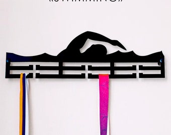 May include: Black metal medal holder shaped like a swimmer in the freestyle stroke. The holder has 10 slots for medals and the text "MEDAL HOLDER <<SWIMMING>>" is printed above the swimmer. Three medals are hanging from the holder, one yellow, one white, and one pink.