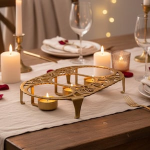 May include: A gold-colored metal candle holder with an ornate floral design, holding several lit tea light candles. The holder sits on a wooden table, with wine glasses and candles in the background, creating a warm, inviting ambiance.