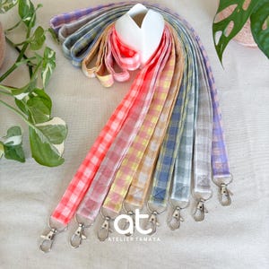 Puede incluir: Una colección de cordones de tela con un patrón de cuadros vichy en varios colores, incluyendo rojo, rosa, amarillo, azul, verde y morado. Cada cordón está equipado con un mosquetón giratorio de metal plateado en el extremo inferior.