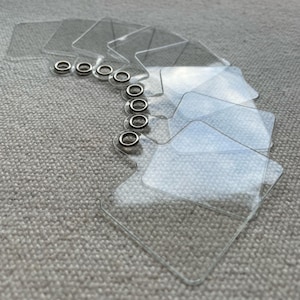 Peut inclure: Un ensemble d'étiquettes carrées en plastique transparent avec des œillets métalliques. Les étiquettes sont disposées en chevauchement, suggérant qu'elles sont conçues pour l'étiquetage ou l'organisation. Les œillets sont argentés et offrent un point de fixation sûr.