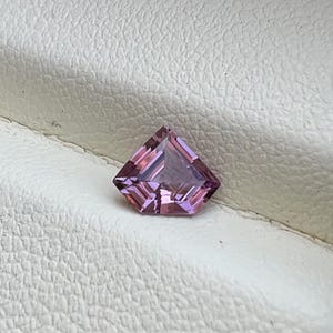 Könnte beinhalten: Ein facettierter, drachenförmiger Edelstein mit einem rosa-violetten Farbton. Der Edelstein liegt auf einer weißen, strukturierten Oberfläche, die seinen geometrischen Schliff und seine Farbe hervorhebt. Die Facetten des Edelsteins reflektieren das Licht und erzeugen einen funkelnden Effekt.