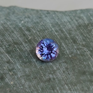 May include: A round, faceted gemstone with a vibrant blue and purple color gradient. The gem is set against a textured, gray-green surface, showcasing its brilliance and clarity. The gemstone's facets reflect light, creating a sparkling effect.