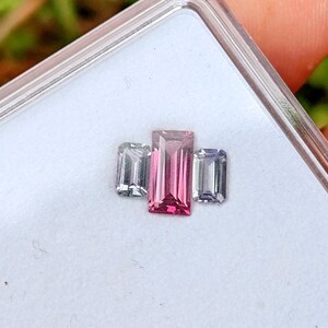 May include: Three rectangular gemstones in a clear plastic case. The center gem is a vibrant pink, flanked by two colorless gems. The gems are set against a white background, with a blurred green background visible.