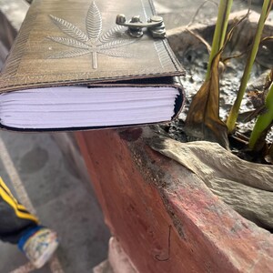 May include: A brown leather journal with a marijuana leaf design embossed on the cover. The journal has a metal clasp and is closed. The journal is sitting on a stone planter.