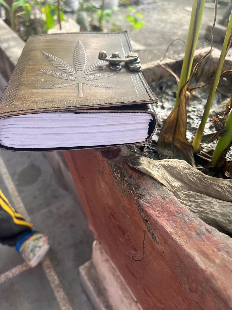 May include: A brown leather journal with a marijuana leaf design embossed on the cover. The journal has a metal clasp and is closed. The journal is sitting on a stone planter.