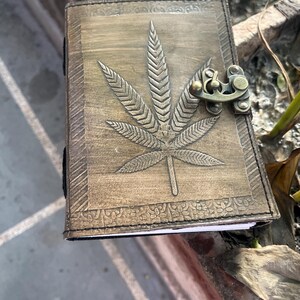 May include: A brown leather journal with a tooled design of a marijuana leaf and a brass clasp.