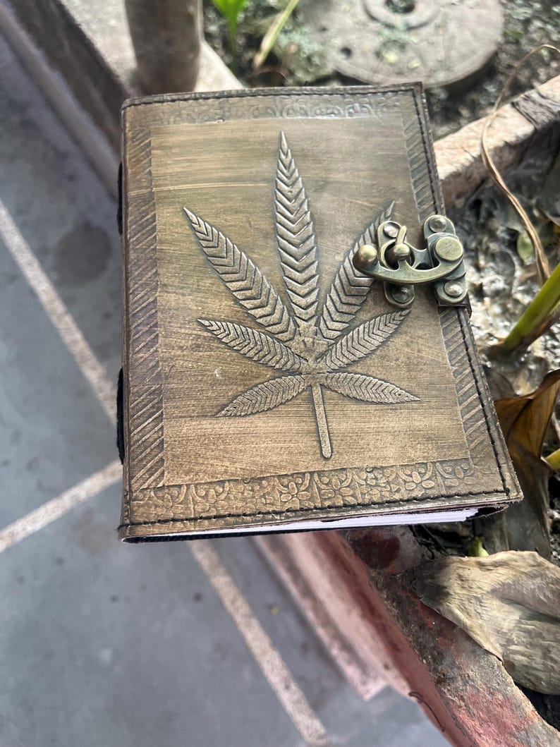 May include: A brown leather journal with a tooled design of a marijuana leaf and a brass clasp.