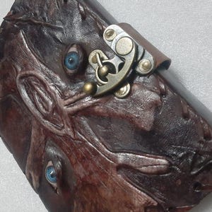 Op de afbeelding: Een bruin leren dagboek met een gedetailleerd reliëf gezichtsontwerp en twee blauwe ogen. Het dagboek heeft een metalen sluiting en een leren riem, wat een unieke en artistieke uitstraling creëert.