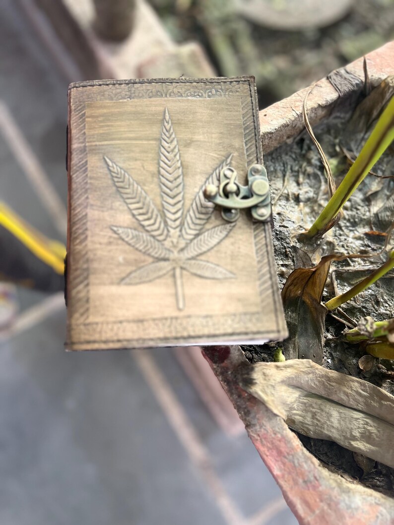 May include: A brown leather journal with a cannabis leaf design embossed on the front cover. The journal has a brass clasp closure.