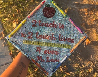 May include: A graduation cap decorated with rhinestones and colorful crayons. The cap features a red apple, a ruler, and the text "2 teach is 2 touch lives 4 ever Ms. Lewis".