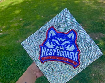 May include: A graduation cap decorated with rhinestones and the West Georgia Wolves logo. The logo is blue, red, and white with a wolf's head in the center.