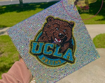May include: A graduation cap covered in iridescent rhinestones. The cap features the UCLA Bruins logo in blue and gold, with a brown bear mascot. The cap is held up in front of a green grassy background.