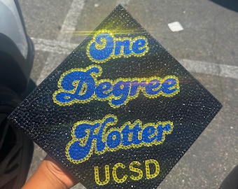 May include: A black graduation cap decorated with blue and yellow rhinestones. The cap says "One Degree Hotter UCSD" in a cursive font.