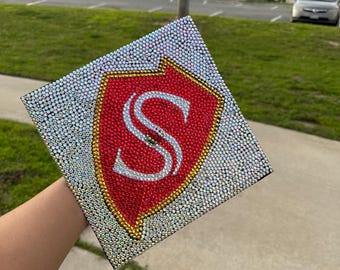 May include: A graduation cap decorated with rhinestones and a red and gold shield with the letter "S" in the center.