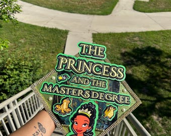 May include: A green graduation cap with the words "The Princess and the Masters Degree" in gold lettering. The cap features a cartoon image of Tiana from Disney's The Princess and the Frog.