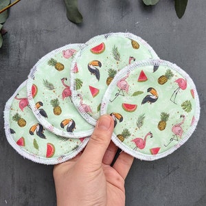 May include: Five round, green cotton makeup remover pads with a white trim. The pads have a tropical print featuring pink flamingos, toucans, pineapples, and watermelon slices.