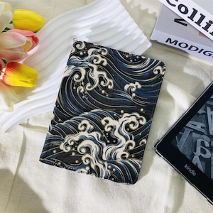 May include: A rectangular case with a wave pattern in navy blue, white, and black. The case is open, revealing a glimpse of a Kindle e-reader. The case is on a white surface with flowers and a book in the background.