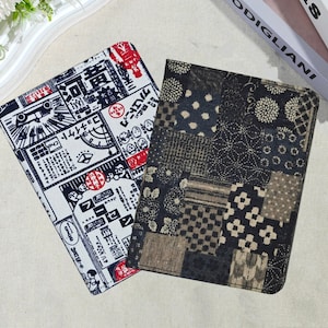 Puede incluir: Dos cuadernos cubiertos de tela. Uno presenta un fondo blanco con un diseño de inspiración japonesa en negro y rojo. El otro tiene un diseño de patchwork en tonos negros, marrones y beige. Los cuadernos son rectangulares y parecen cerrados.