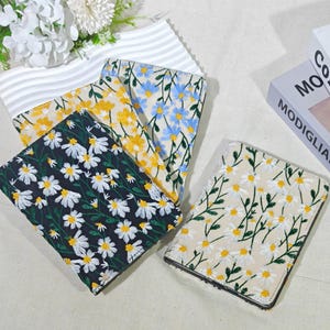 May include: A collection of floral embroidered passport holders in various colors. Designs include white daisies on a black background, yellow daisies on a cream background, and blue and yellow flowers on a white background. A white and gray striped fabric is also visible.