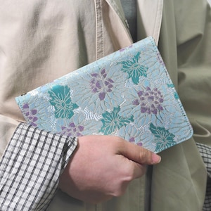 May include: A floral patterned book cover in shades of light blue, teal, and purple. The cover features a repeating pattern of stylized flowers. The cover is held in a person's hand, with a tan coat and plaid shirt visible in the background.