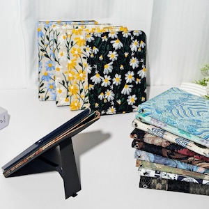 Puede incluir: Varias fundas para tabletas con bordados florales, incluyendo margaritas, en azul, amarillo y blanco. Un soporte negro sostiene una tableta, y también se ve una pila de telas estampadas. Se ofrecen diversos diseños florales.