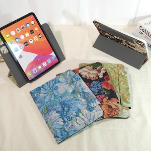 以下が含まれることがあります： グレーのスタンドにiPadが表示され、画面が見えています。青、黒、緑のデザインの、花柄のiPadケースがいくつか見えます。ケースはテクスチャード加工された生地で作られています。