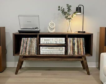 May include: A mid-century modern style entertainment center with a turntable, stereo equipment, and vinyl records. Two tall speakers flank the center. A black desk lamp and decorative vase are on top of the console. The room has white sofas and light wood flooring.