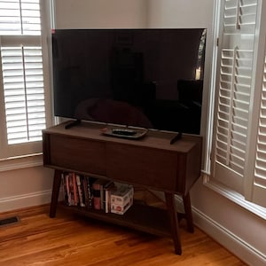 May include: A large flat-screen television with a black display sits atop a dark brown wooden media console. The console features a rectangular cabinet with two doors and an open lower shelf containing several books and a small white box.