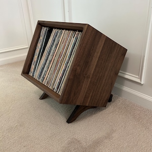 May include: A dark brown wooden record storage crate with a slanted open front, designed to hold a collection of vinyl records upright. The crate features a boxy frame supported by two angled wooden legs, showcasing a natural wood grain finish.