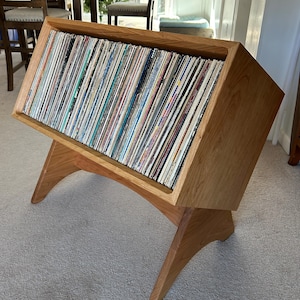 May include: A wooden record storage unit with a slanted top, holding numerous vinyl records. The unit is made of light-colored wood and has a unique, angled design with angled legs. The records are visible, showcasing various album covers.