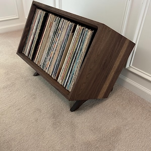 May include: A dark brown wooden vinyl record storage console with an angled front opening. The unit features a rectangular frame with three internal dividers, holding a large collection of vinyl records with various colorful album covers.