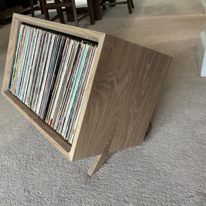May include: Angled wooden record storage unit filled with vinyl records. The unit is made of light-colored wood and has a slanted design. The records are visible inside the open compartment, showcasing a collection of album covers.