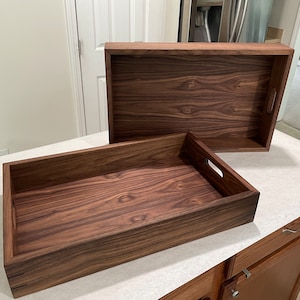 May include: Two wooden trays made of walnut wood. The trays are rectangular and have a natural wood finish. The larger tray is sitting on top of the smaller tray.