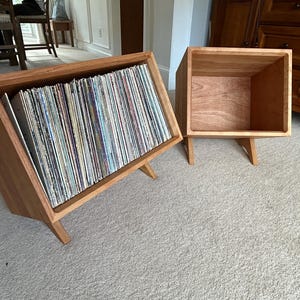 May include: Two wooden record storage units with angled sides and legs. One unit is full of vinyl records, while the other is empty.