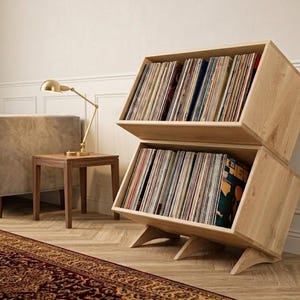 May include: A light-colored wooden storage unit consisting of two stacked, angled open-front boxes designed to hold vinyl records. The unit features a natural wood grain finish and is supported by three curved wooden legs at the base.
