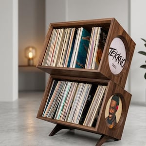 May include: A wooden record storage unit with two angled compartments. Each compartment holds numerous vinyl records. The unit has a dark wood finish and a circular design element with the word "TEKKNO" on one side and a portrait on the other.