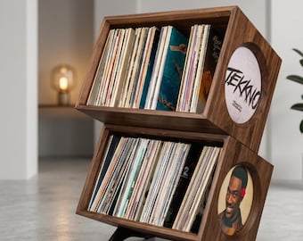 May include: A wooden record storage unit with two angled compartments. Each compartment holds numerous vinyl records. The unit has a dark wood finish and a circular design element with the word "TEKKNO" on one side and a portrait on the other.