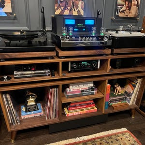 May include: A walnut wood media console featuring open shelving filled with vinyl records, books, and a gold award trophy. The top surface holds two turntables and a McIntosh amplifier with glowing blue meters and vacuum tubes on display.