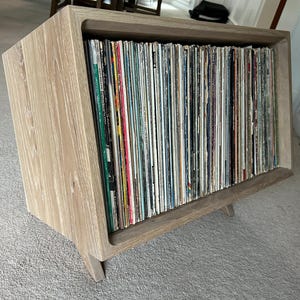 May include: A light-colored wooden record storage cabinet with a slanted front. The cabinet is filled with vinyl records. The cabinet has a simple, modern design with angled legs and a recessed front.