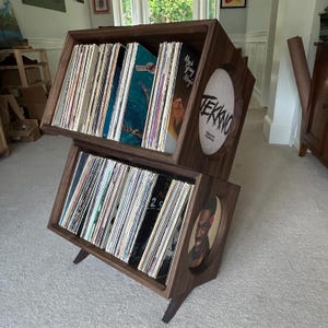 May include: A two-tiered dark wood vinyl record storage stand with angled shelves. Each tier holds multiple records in sleeves. The side panels feature circular cutouts, with one displaying the text TEKKNO and the other a portrait of a person.
