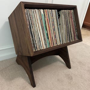 May include: A dark brown wooden record storage cabinet with a slanted top and curved legs. The cabinet is filled with vinyl records. The cabinet is made of solid wood and has a modern design.
