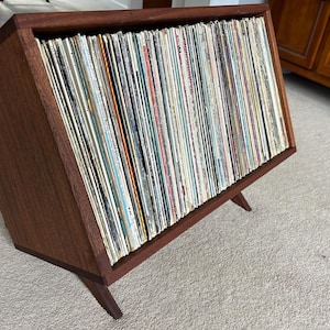 May include: A wooden record storage unit with a slanted front and angled legs. The unit is filled with vinyl records.