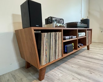May include: A mid-century modern style media console made of wood, with angled sides and four legs. The console features open shelving for vinyl records, gaming consoles, and a turntable. Two black speakers sit on top.