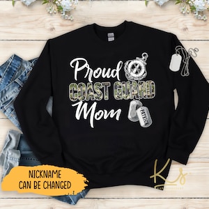 May include: Black sweatshirt with a camouflage print and the text "Proud Coast Guard Mom" in white. A silver charm with an anchor is above the text. A silver dog tag with the name "Patrick" is below the text. The text "NICKNAME CAN BE CHANGED" is in yellow at the bottom of the sweatshirt.