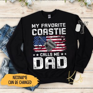 May include: Black long-sleeve sweatshirt with a graphic design featuring an American flag, a dog tag with the name "Joshua" on it, and the text "My Favorite Coastle Calls Me Dad".